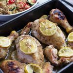 Horizontal image of chicken pieces in a dark pan topped with lemons, next to a quinoa and vegetable side dish.
