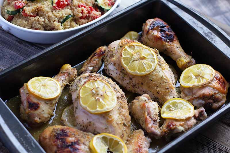 Horizontal image of chicken pieces in a dark pan topped with lemons, next to a quinoa and vegetable side dish.