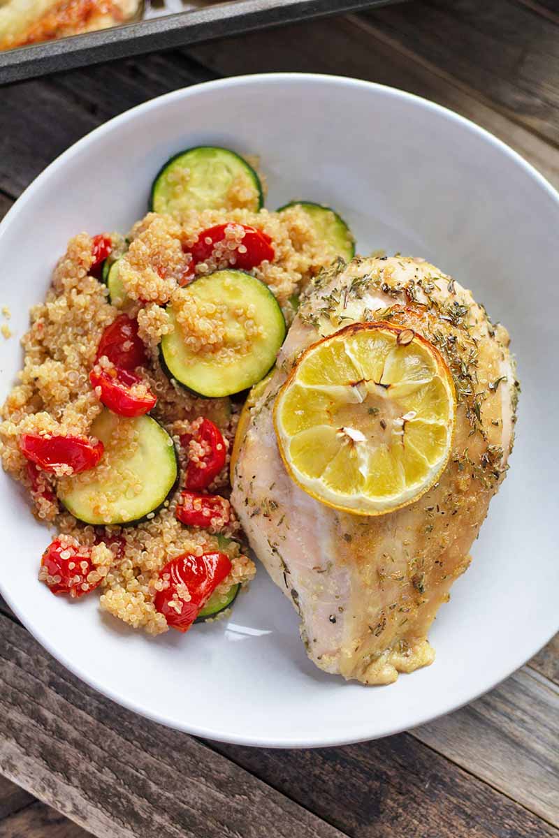 Vertical top-down image of a white plate with a poultry breast topped with a thin slice of lemon, next to a vegetable quinoa salad.