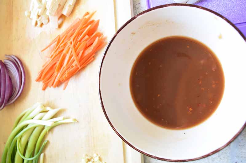 Horizontal image of a bowl with a brown sauce next to prepped vegetables.