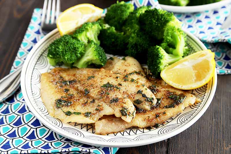 Horizontal image of a plate with cooked sole with herbs on a plate with broccoli and lemons, with a blue napkin and fork.