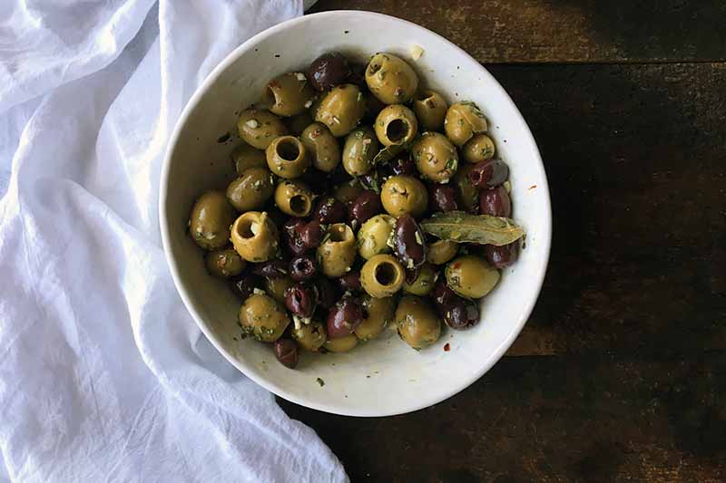 Horizontal image of a large white bowl with a mixture of garlic, herbs, and olives.