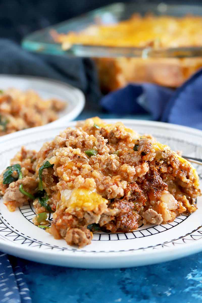 Vertical image of two plates with a cheese and quinoa bake, with the main casserole dish in the background on a blue napkin.