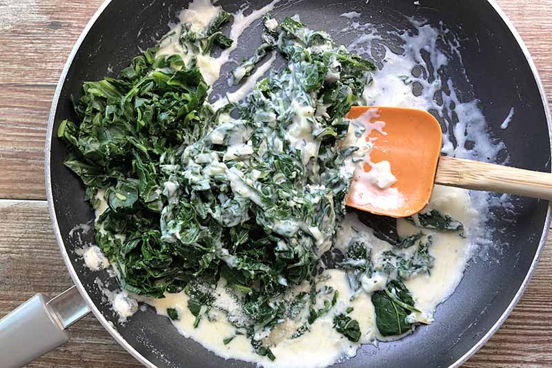 Overhead closely cropped image of cooked collard greens in a cream sauce being stirred with an orange rubber spatula with a wooden handle in a large nonstick frying pan, on a light brown wood surface.