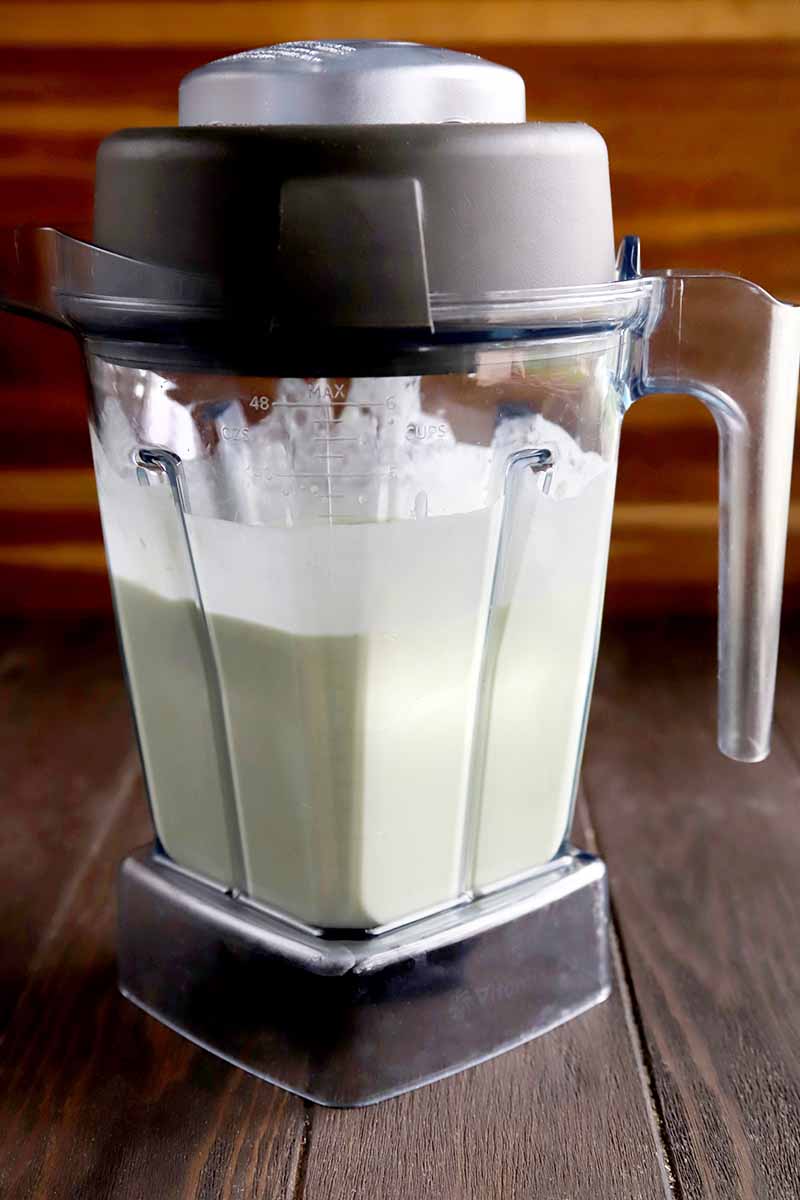 Vertical image of a pale green smoothie in a clear plastic Vitamix pitcher-style blender container with a black lid, on a brown wood surface against a striped lighter brown background.