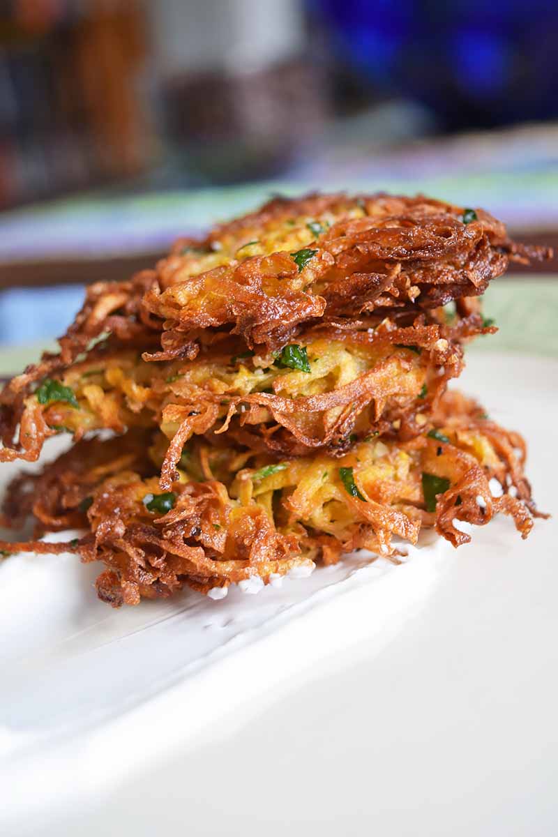 Vertical image of a stack of three fried golden brown latkes, on a white surface with blue and brown objects in soft focus in the background.