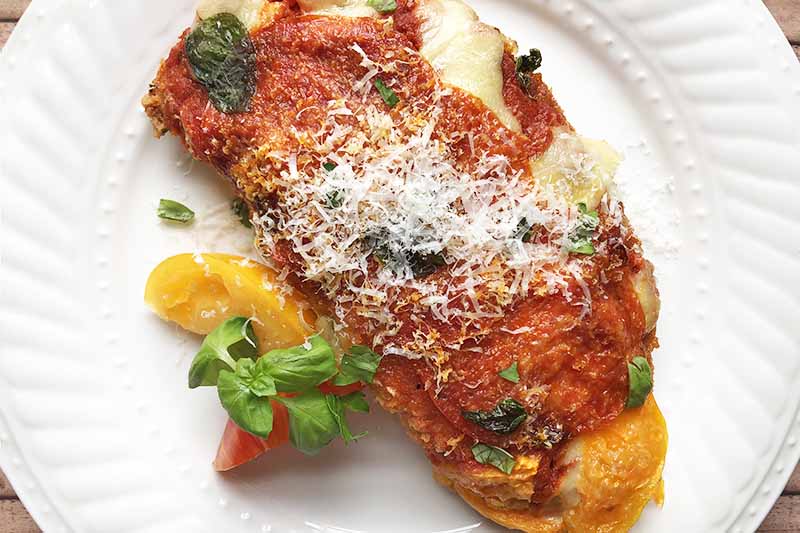 Overhead horizontal image of chicken parmesan with pepperoni on a white plate, garnished with slices of red and yellow tomato and a sprig of green basil, sprinkled with grated hard Italian cheese.