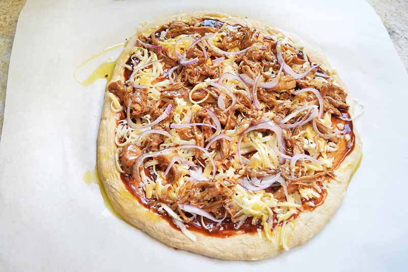 Horizontal overhead image of a small barbecue chicken pizza on a white parchment paper surface, drizzled with olive oil and ready to be baked.
