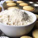Horizontal image of a small bowl filled with a light white crumbly powder next to mini cupcakes.