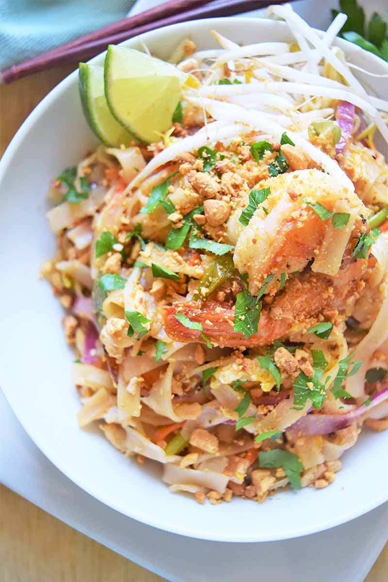 Vertical image of shrimp pad Thai in a white bowl next to chopsticks and a blue napkin.