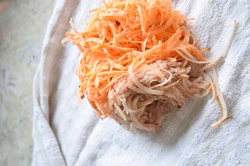 Overhead horizontal image of a finely shredded orange and white potato that has been squeezed to remove excess moisture, on a white cloth on top of a gray speckled surface.