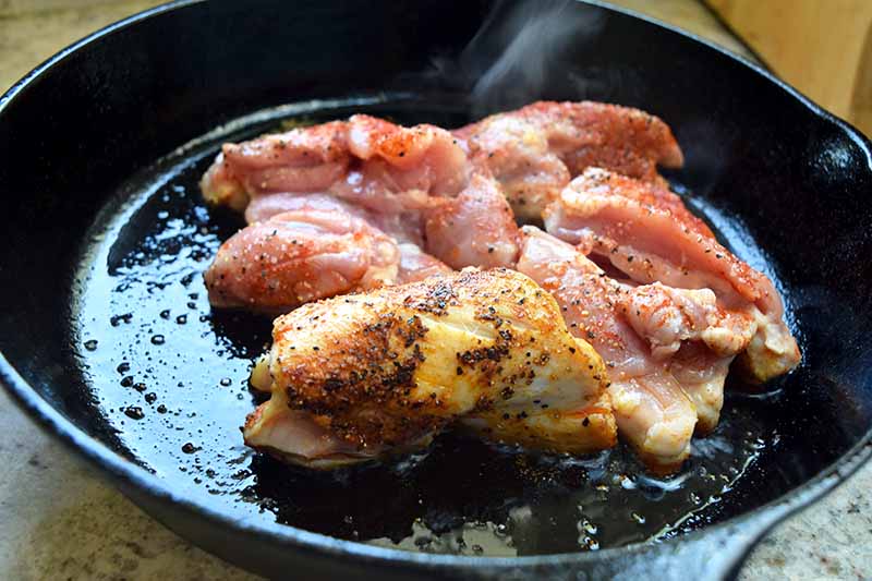 Horizontal image of chicken thighs being fried until golden brown in a large pan.
