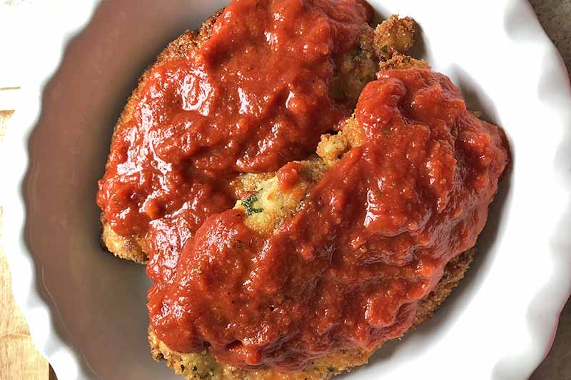 Horizontal overhead image of two breaded chicken breasts in a white ceramic pie dish with a fluted edge, topped with marinara sauce.