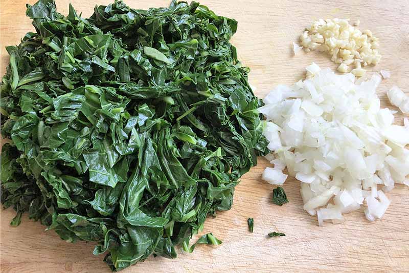 Overhead horizontal image of piles of chopped collard greens, chopped onion, and minced garlic, on a pale beige unfinished wood cutting board.