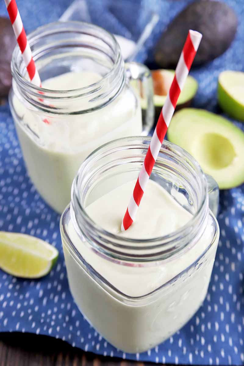 Vertical oblique overhead image of two glass jars with handles filled with a homemade smoothie, with white and red striped paper straws in each, on a blue cloth with white spots, with lime wedges and cut halves of fresh avocado.
