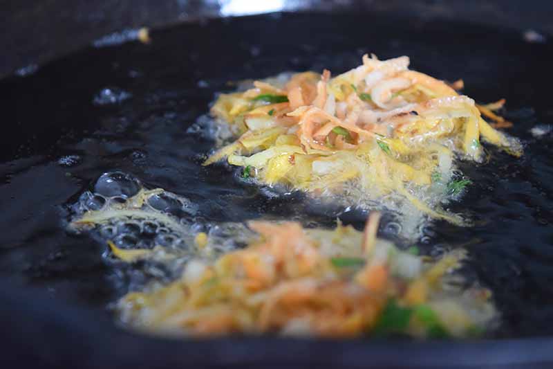 Horizontal image of shredded root vegetable pancakes frying in a large pan of hot vegetable oil.
