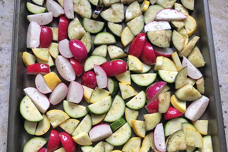 Horizontal overhead image of a baking sheet filled with sliced yellow squash, zucchini, and radishes.