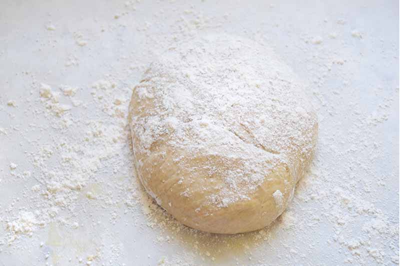 Overhead horizontal image of a ball of lightly floured pizza dough on a white surface.