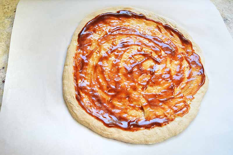 Overhead horizontal image of a round of pizza dough with barbecue sauce ladled and spread in a circular pattern on top, on a piece of white parchment paper, on a beige kitchen countertop.