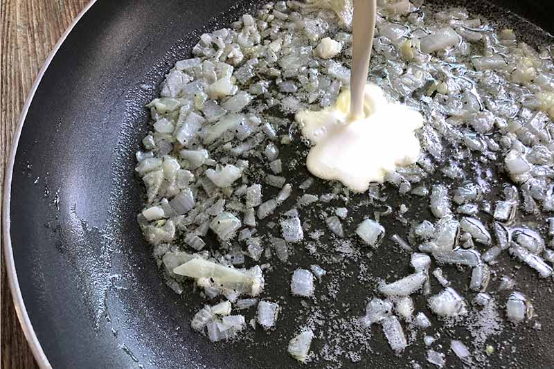 Horizontal image of cream being poured into a pan of sauteeing minced garlic and chopped onion in oil, in a nonstick frying pan, on a wood surface.