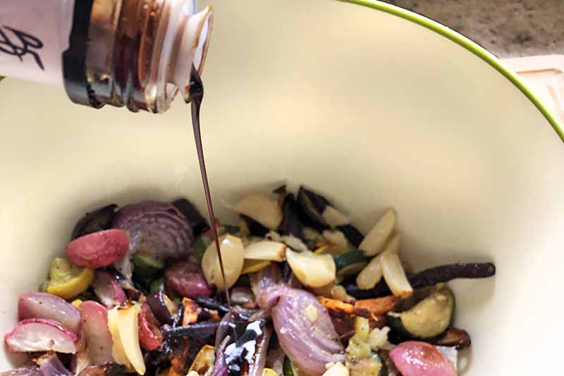 Closely cropped horizontal image of balsamic vinegar being poured from a bottle into the bowl of vegetables below.