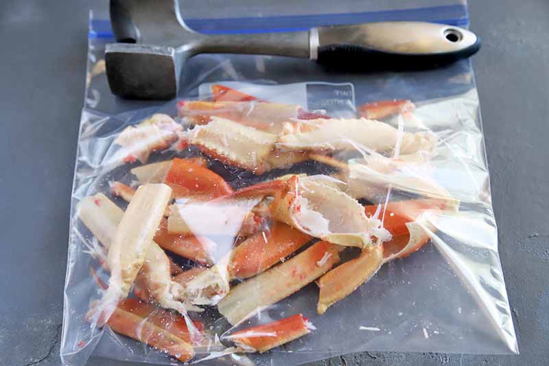 Horizontal image of an airtight bag filled with crab legs next to a meat mallet.