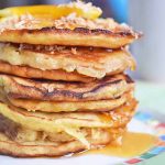 Horizontal image of a stall stack of pancakes on a colorful plate with syrup, orange slices, and coconut flakes.