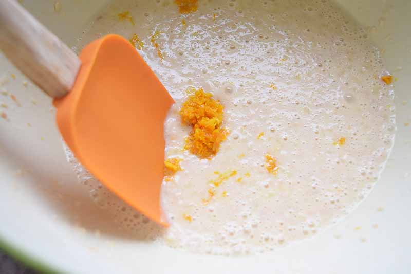 Horizontal image of a spatula mixing together orange zest in a light batter.