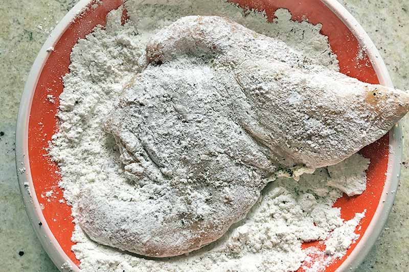 Horizontal overhead image of a chicken cutlet being dredged in flour, in an orange and white ceramic bowl on a speckled beige countertop.