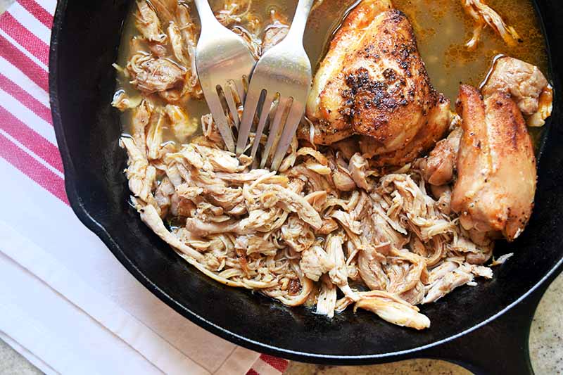 Two forks in a cast iron pan of cooked chicken thighs, about half of which have been shredded, on a beige surface with a white and red striped cloth kitchen towel.