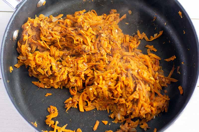 Horizontal overhead image of shredded sweet potato coated with spiced, sauteeing in a large nonstick frying pan, on a white painted wood surface.