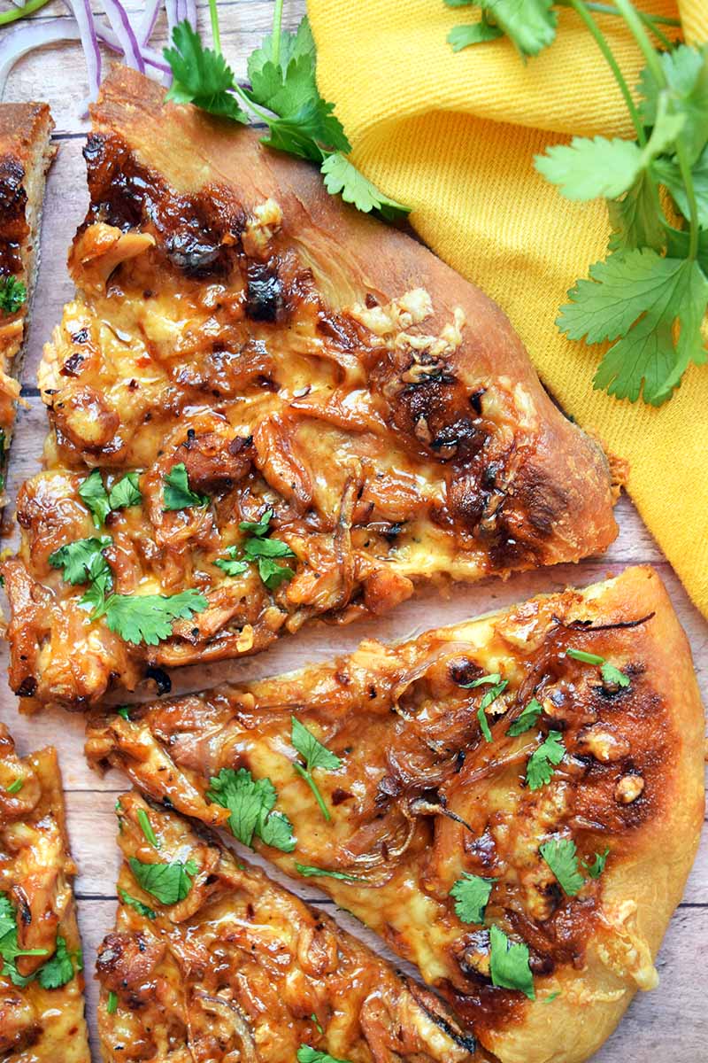 Vertical closely cropped overhead image of slices of homemade barbecue chicken pizza topped with fresh cilantro, on a wood surface with a yellow cloth and scattered sprigs of fresh herbs.