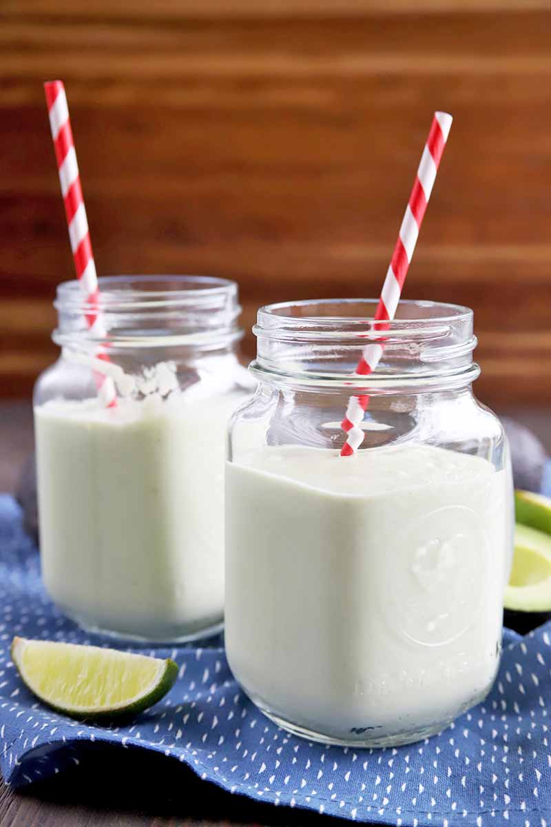 Vertical image of two glass jars with handles filled with a homemade smoothie that has a pale green color, with red and white striped straws in each, on a blue cloth with white spots, with scattered lime wedges and cut avocado, against a brown striped background.