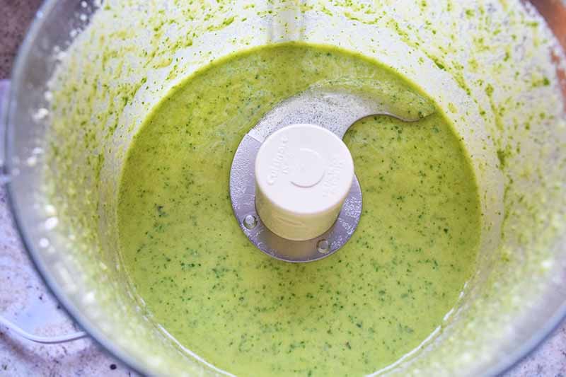 Horizontal closely cropped overhead image of pale green blended herb dressing in a food processor with a clear plastic canister.
