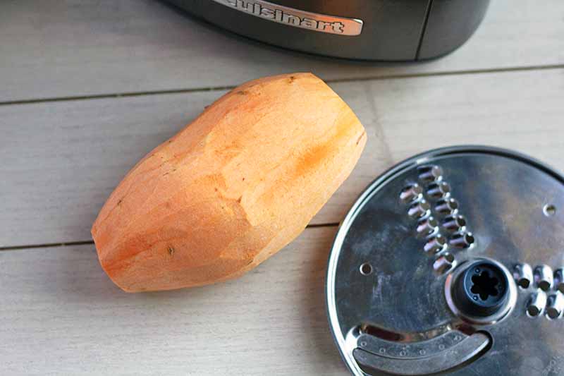 Horizontal overhead image of a peeled orange sweet potato with a metal food processor shredding disc and the black plastic base of the appliance, on a white wood surface with three slats.