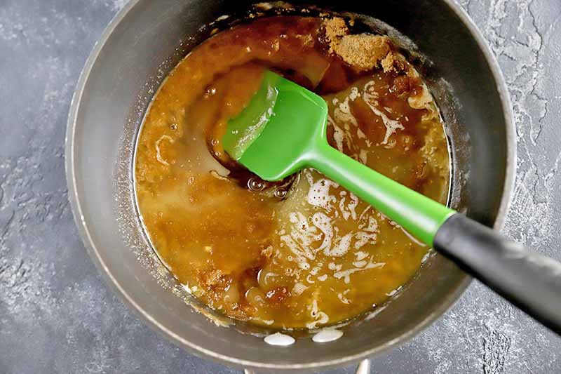 Horizontal overhead image of a green and black rubber spatula stirring brown sugar into a caramel mixture in a saucepan, on a gray surface.