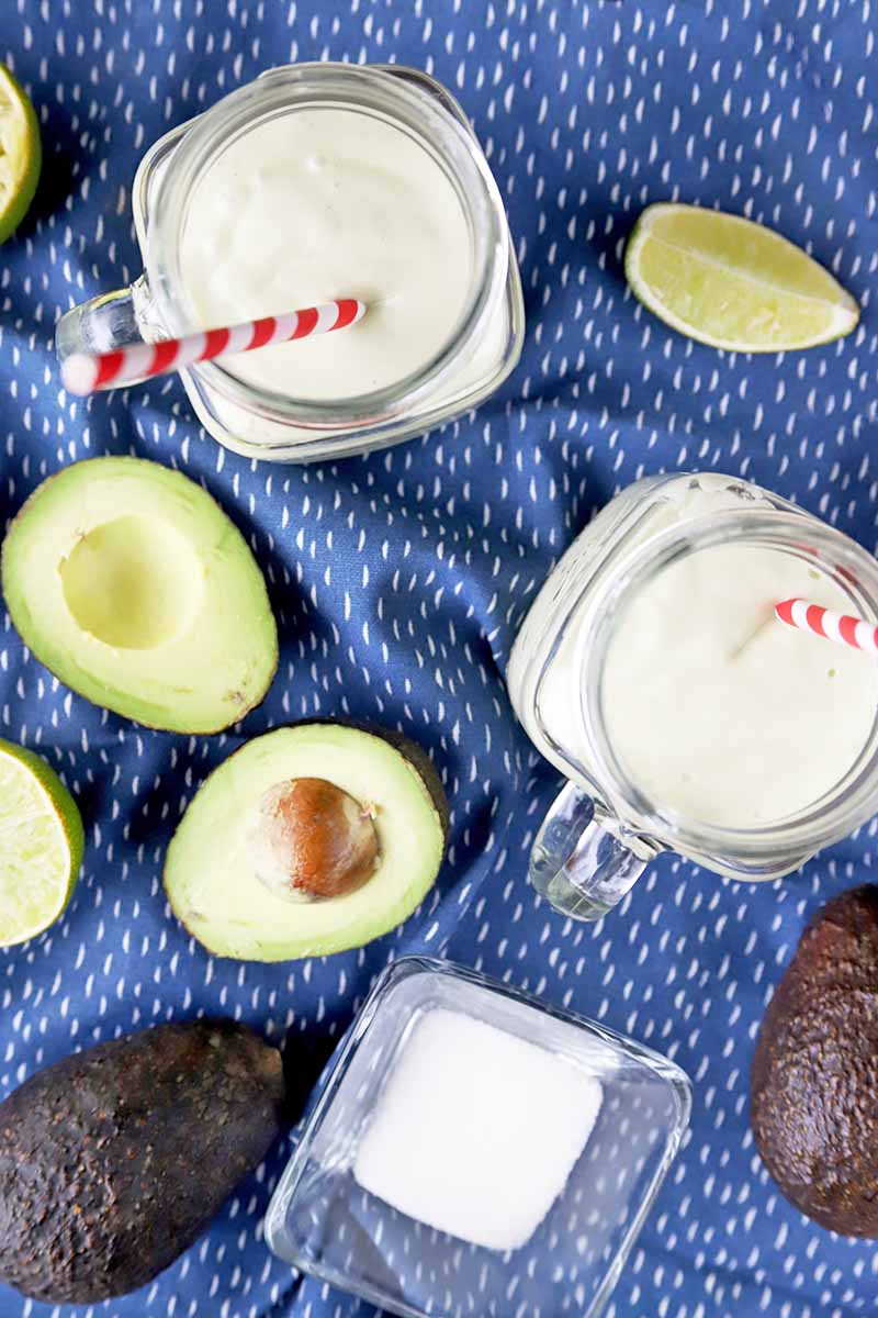 Vertical overhead image of a white smoothie in two glass jars with handles with red and white paper straws inserted in each, on a blue cloth with white speckles, with whole and halved avocados and lime wedges, and a square glass bowl of coconut milk.