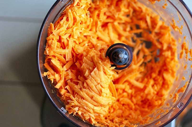Horizontal closely cropped overhead image of shredded orange sweet potato in a food processor, on a gray surface.