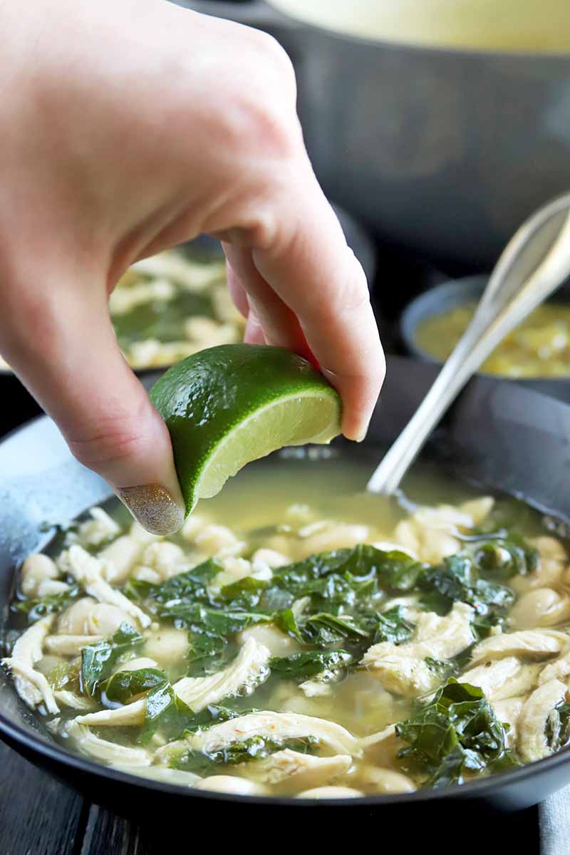 A hand squeezes a wedge of lime over a black bowl of chicken, kale, and cannellini bean soup with a spoon sicking out of it, on a dark brown surface with an enameled gray and creme-colored cooking pot in the background.