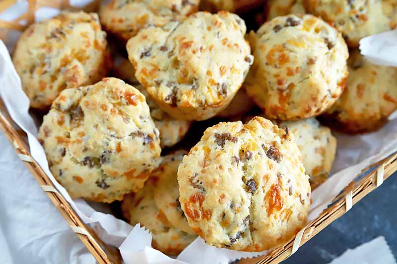 Horizontal closely cropped oblique overhead image of a basket lined with parchment paper and filled with sausage cheese biscuits, on a blue-gray surface topped partially with more paper.