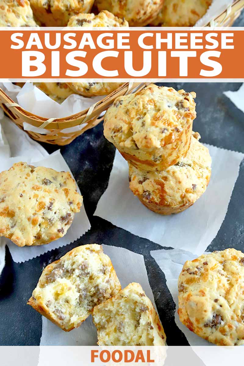 Vertical oblique overhead image of sausage cheese biscuits in a basket, and stacked and scattered on a gray surface with squares of white parchment paper, with one at the bottom of the frame that has been torn in half to show the interior crumb, printed with orange and white text near the top and at the bottom of the frame.