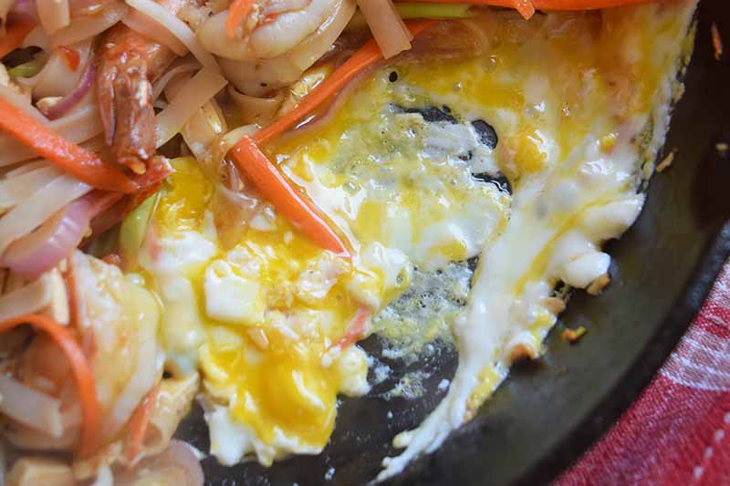 Horizontal image of scrambling an egg in a dish with vegetables, noodles, and seafood.