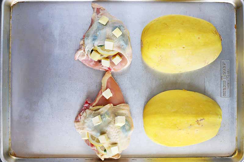 Overhead image of raw split chicken breasts with cubes of butter on top of the skin, and two pieces of halved yellow spaghetti squash arranged cut side down on a baking sheet.