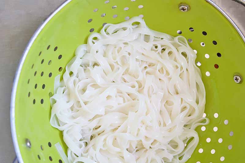 Horizontal image of a green colander with soaked rice noodles.