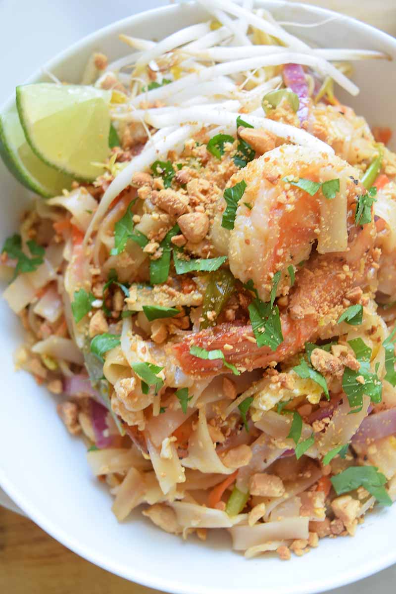 Vertical top-down image of a noodle dish with seafood, fresh herbs, nuts, bean sprouts, and lime wedges.