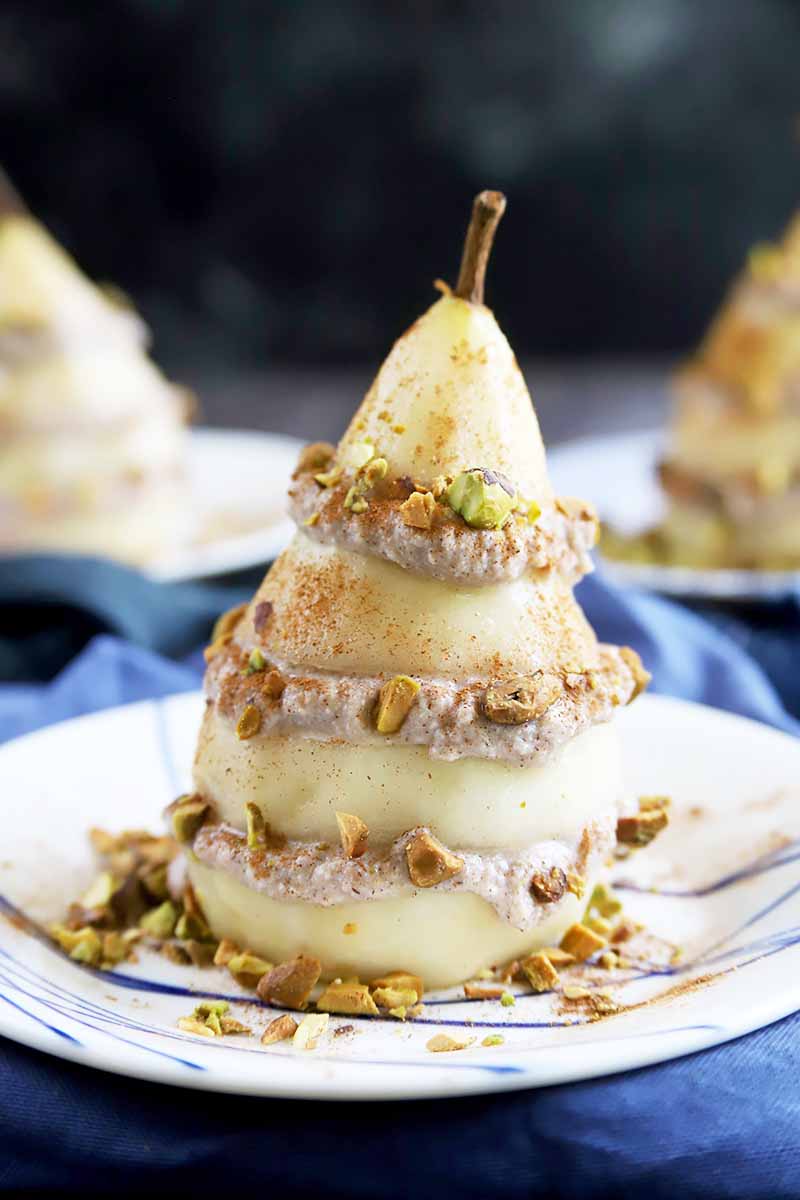 Vertical image of a pear and ricotta stack covered in chopped pistachios on a plate on a blue napkin.