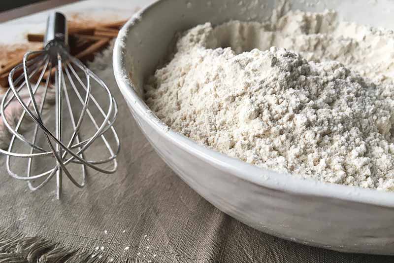 Horizontal image of a white bowl filled with a flour and spice mixture next to a whisk, on a gray cloth with fringe and cinnamon sticks in soft focus in the background.