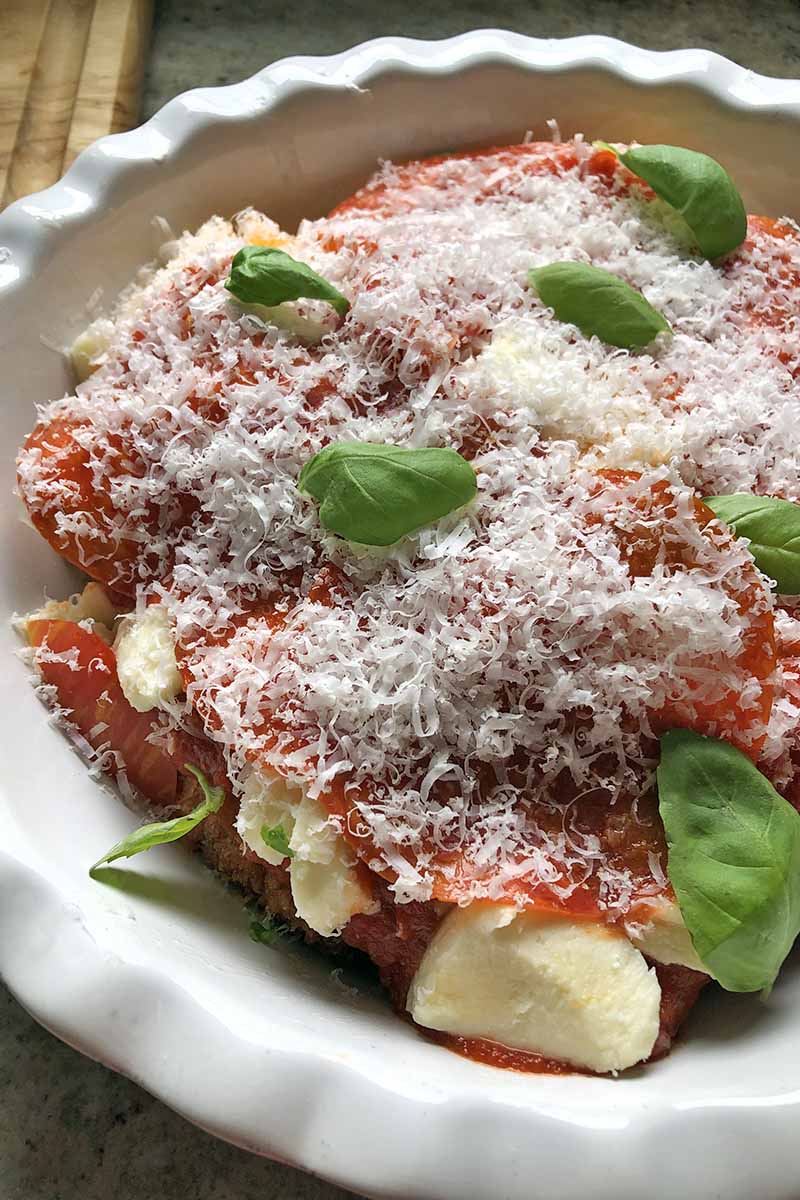 Vertical oblique overhead image of chicken parmesan in a white ceramic pie plate, topped with mozzarella, marinara sauce, grated Parmigiano-Reggiano cheese, and torn leaves of fresh basil.