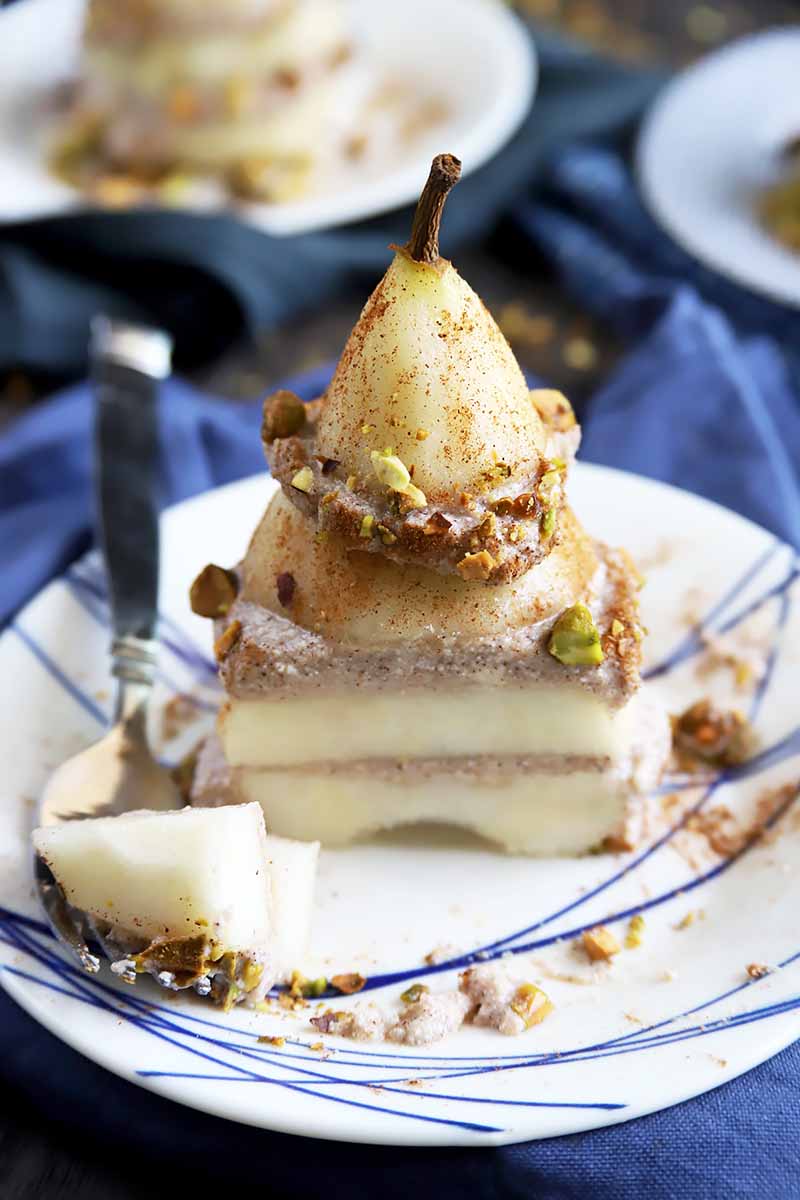 Vertical image of a layered dessert with pear and spiced ricotta filling with a piece removed on a plate garnished with chopped nuts.