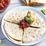 Horizontal overhead image of a quesadilla on a white plate that has been cut into quarters and topped with dollops of guacamole and salsa, on a white surface with a glass dish of sauce, another filled tortilla that has been sliced and stacked, and three wedges of lime.